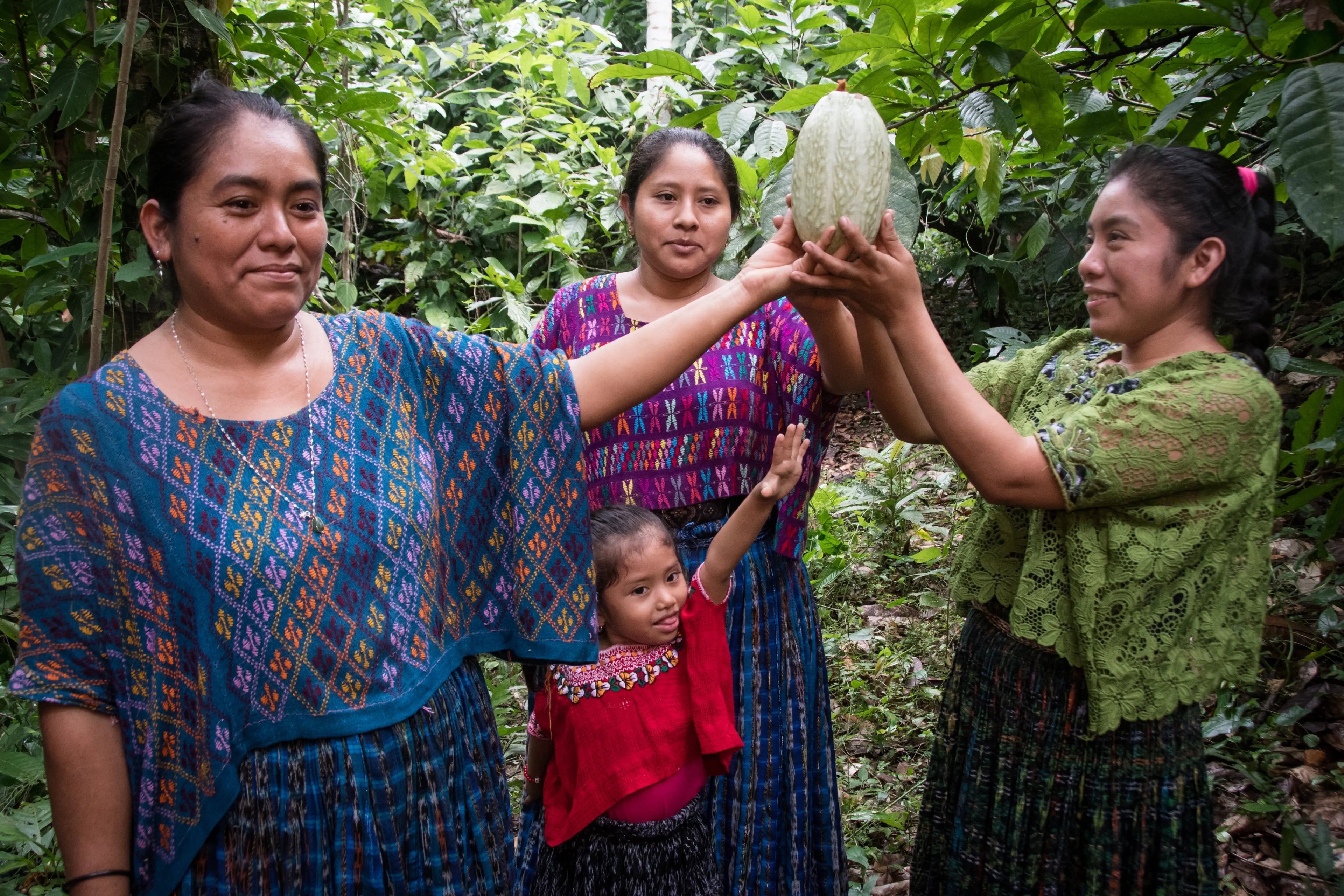 Drei Maya-Frauen und ein Maya-Mädchen stehen im Dschungel und halten eine Kakao-Schote