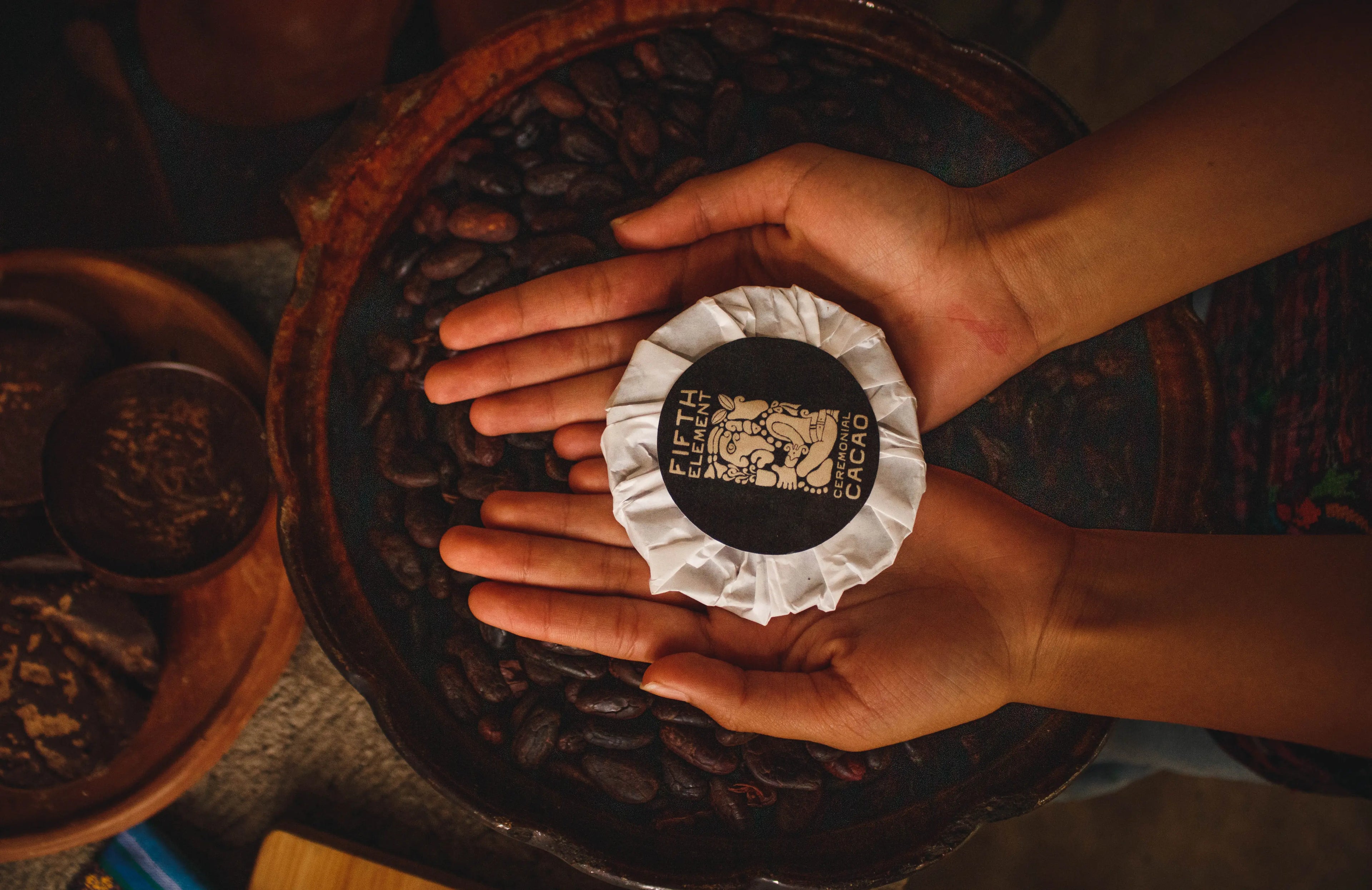 Hands holding a packed ceremonial cacao above a clay bowl filled with roasted cacao beans